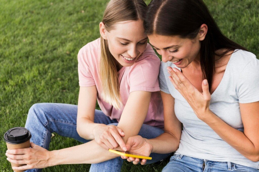 Two female friends sitting on grass, engaging with a smartphone, sharing laughter and conversation, with a coffee cup nearby, emphasizing outdoor social interactions.